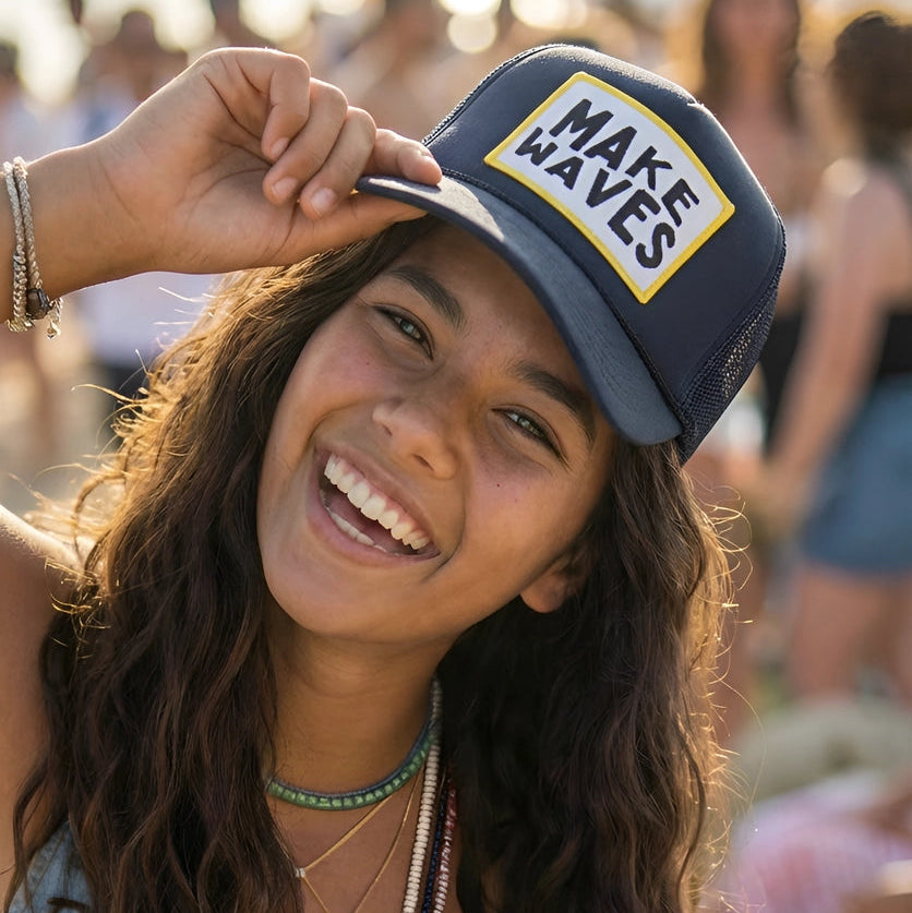 Woman wearing a 'Make Waves' cap on a beach with blurred people in the background