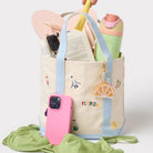 Beige tote bag with colorful handles, pink phone case, and green fabric on a light gray background